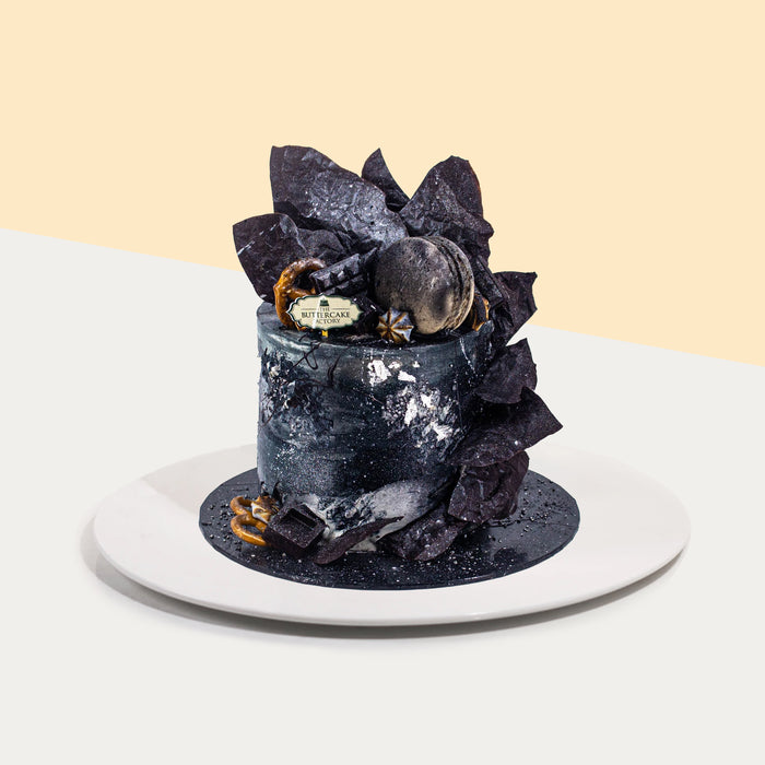 Black buttercream cake with touches of silver edible spray, topped with a macaron, chocolate shards and pretzels