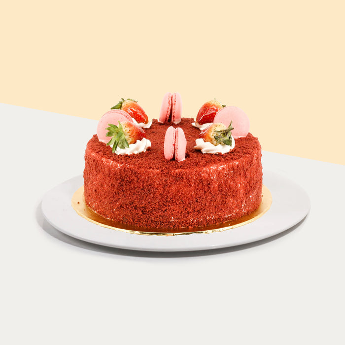 Red velvet cake covered in cream cheese and red velvet crumbs, topped with strawberries and macarons